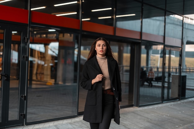 Fashion model pretty stylish young woman in a fashionable black coat in a beige shirt walks near a vintage glass building in the city. Beautiful modern girl in elegant clothes outdoors. Trendy look.