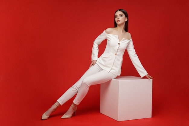 Fashion model sits on cube in white suit pants Beautiful young woman Studio shot Red background