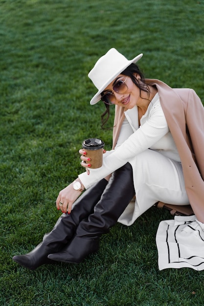 Fashion model with cup of coffee seat on the grass. Attractive woman weared whihe hat, dress, jacket and sunglasses.