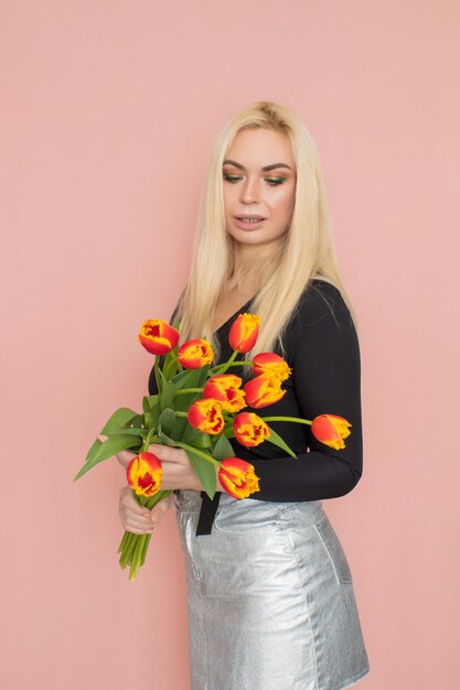 Fashion model woman in fashionable clothes on pink background wearing stylish clothing black blouse silver skirt posing in studio holding red tulips in her hands