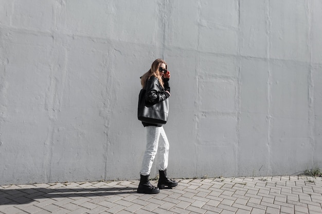 Fashion model woman in stylish casual clothes with leather jacket jeans boots and black bag puts on a sunglasses and walks near a gray wall on the street