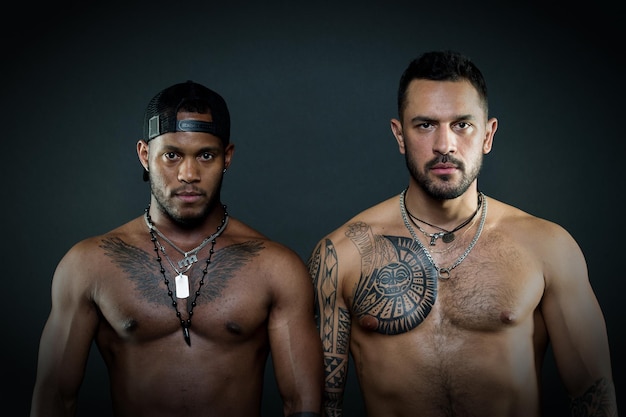 Fashion models with serious faces and fit bodies isolated on black background. Caucasian man with geometrical tattoos standing next to African guy with eagle tattoo on chest wearing cap backwards.