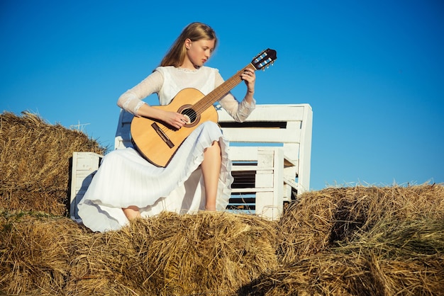 Fashion musician in white dress on sunny nature. Woman guitarist perform music concert. Albino girl hold acoustic guitar, string instrument. Sensual woman play guitar on wooden bench