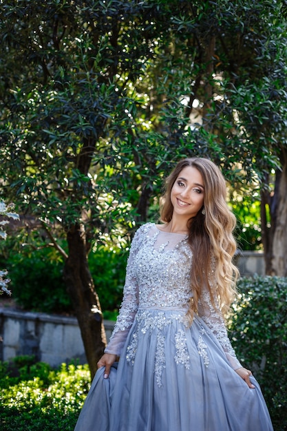 Модные наружные фото элегантной красивой женщины со светлыми волосами в роскошном платье с блестками и серебряными аксессуарами, позирующей в летнем парке