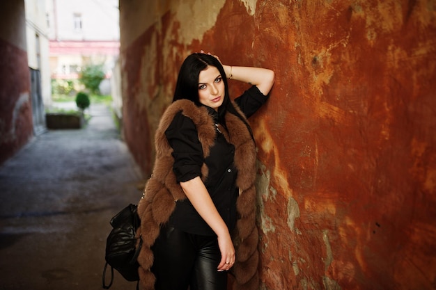 Fashion outdoor photo of gorgeous sensual woman with dark hair in elegant clothes and luxurious fur coat at old street with grunge walls