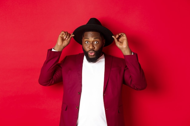 Fashion and party concept. handsome black man wearing hat going on holiday celebration, looking excited, standing over red background