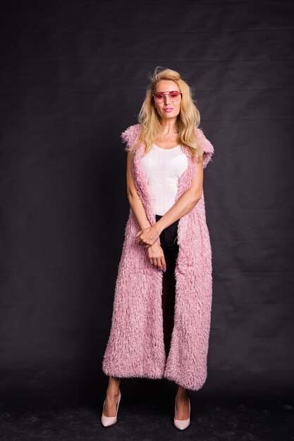 Fashion and people concept - young blonde woman in glasses standing in pink fur coat and smiling.