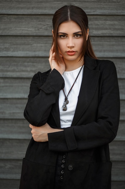 Fashion portrait of a beautiful model woman in a black stylish coat near a wooden wall outdoors