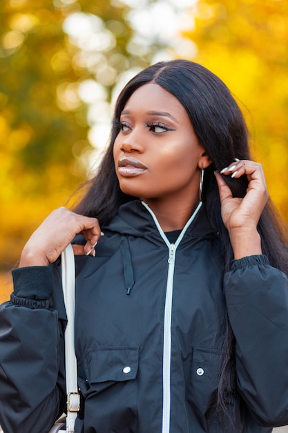 Fashion portrait of a beautiful young African American woman in a stylish black jacket with a handbag straightens her hair and walks in the park against a background of colorful yellow autumn foliage