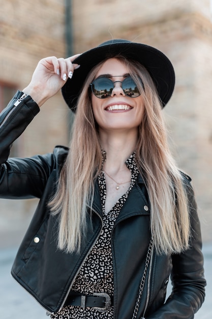 Fashion portrait of a beautiful young happy woman in a vintage dress with a leather jacket and stylish sunglasses walks on the street on a sunny day