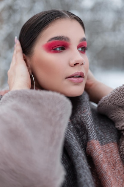 Fashion portrait of a beautiful young stylish woman with bright pink makeup in winter fashionable clothes with a scarf walks on a winter day in a snowy park