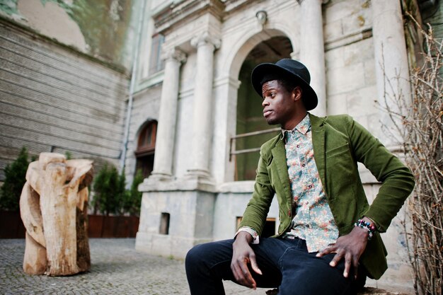 Fashion portrait of black african american man on green velvet jacket and black hat sitting at felled tree background old vintage house with arch and columns