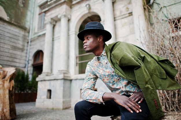 Fashion portrait of black african american man on green velvet jacket at his shoulders and black hat sitting at felled tree background old vintage house with arch and columns