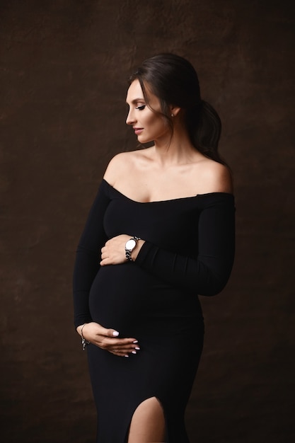 Fashion portrait of a gorgeous pregnant model girl wearing black evening dress posing over brown bac...