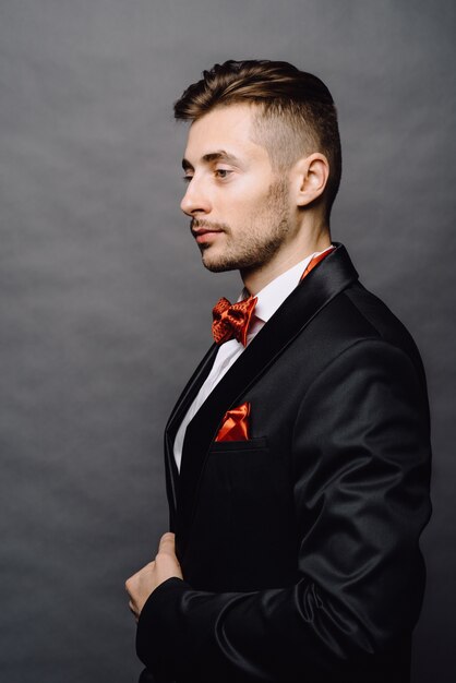 Fashion portrait of handsome elegant man with curly hair wearing tuxedo posing on gray background in studio