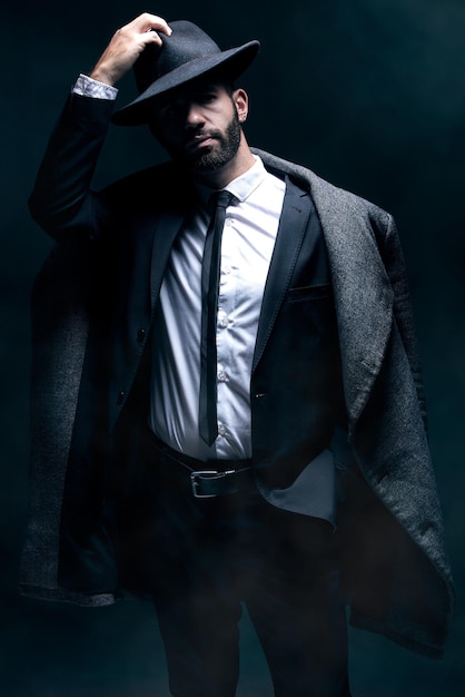 Модный портрет и мужчина в темной студии с уверенной одеждой и шляпой в винтажном стиле Модельный человек в костюме и пальто для зимнего бизнеса или тайны в роли шпиона на черном фоне