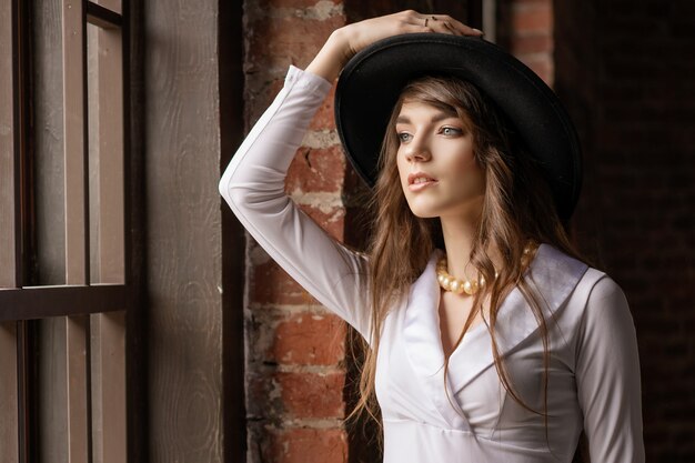 Фасонируйте портрет шляпы молодой красивой уверенно женщины нося, представляя внутри на окне