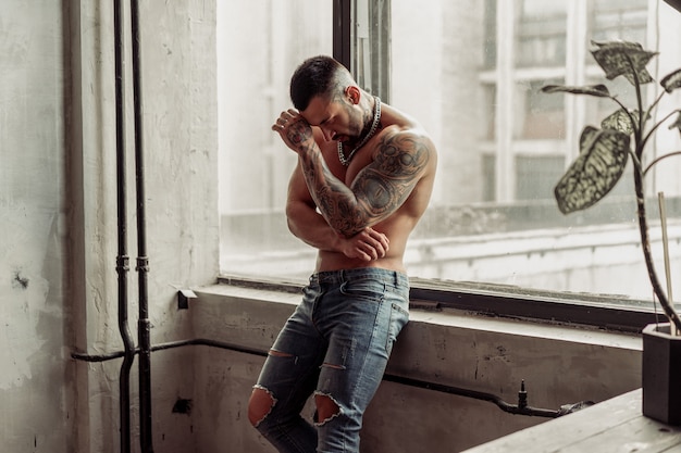 Fashion portrait of Sexy naked male model with tattoo and a black beard standing in hot pose on near the window. Loft room interior with grey concrete wall. 