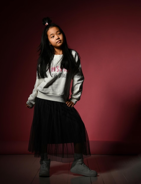 Fashion portrait of a small korean model girl posing\
professionally against a red background girl in a black skirt white\
sweater sneakers children\'s fashion concept