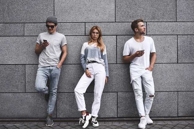 Foto ritratto di moda di tre migliori amici in posa in strada, indossando abiti eleganti e jeans contro il muro grigio.