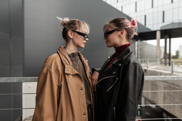 Fashion portrait of two beautiful young women in stylish leather jacket with sunglasses looking at each other in the city