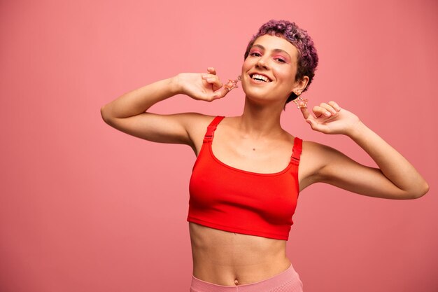 Fashion portrait of a woman with a short haircut of purple color and a smile with teeth in a red top on a pink background happiness High quality photo