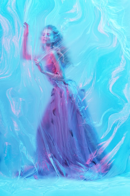 fashion portrait of young beautiful woman under polyethylene. blond curly hair, makeup, fluffy dress with feathers, blue and red colors. art photo