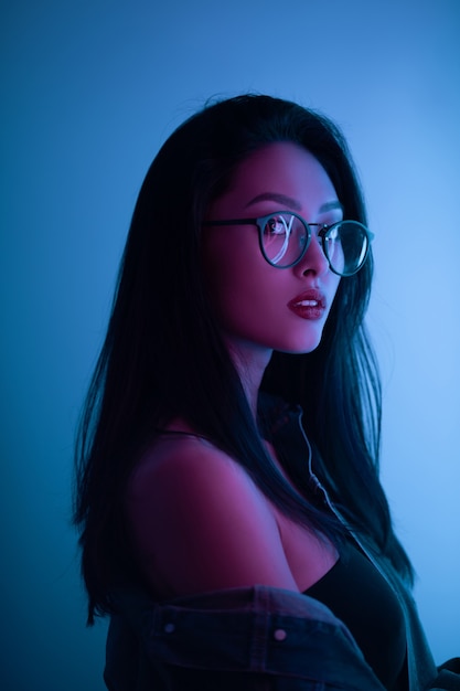 Fashion portrait of young elegant woman. Young asian millennial girl in fashion style wearing glasses casual clothes, neon light blue background, night life and fashion lifestyle.