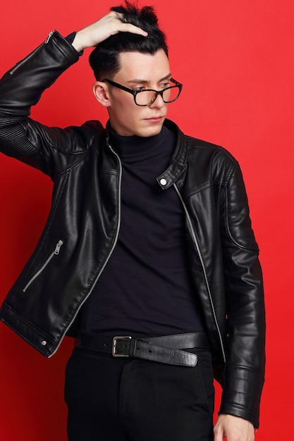若いハンサムな男のファッションの肖像画。黒革のジャケット、タートルネック、メガネ、ブルネットの髪、赤い背景