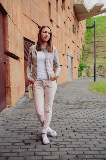 Fashion portrait of young trendy woman wearing gray jacket, pink jeans and white satchel bag over a stone wall