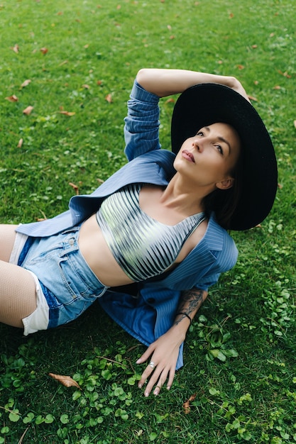 파란색 줄무늬 재킷과 검은 모자를 입은 젊은 여자의 패션 초상화. 여자는 도시에있는 공원에서 녹색 잔디밭에 누워