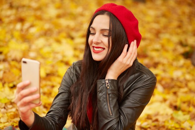 Fashion portrait of young woman outdoor in autumn park or foreest, lady wearing lather jacket, and red beret