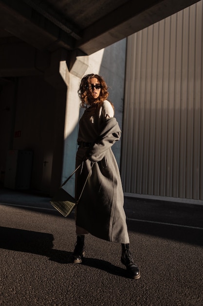 Фото Модная модель довольно кудрявой женщины с солнцезащитными очками в стильном длинном пальто и кожаной сумке гуляет по улице. городской женский стиль casual