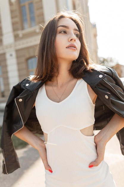 Fashion pretty girl model with beautiful face in fashionable white dress with black leather jacket walks in the city urban casual woman's style clothes look