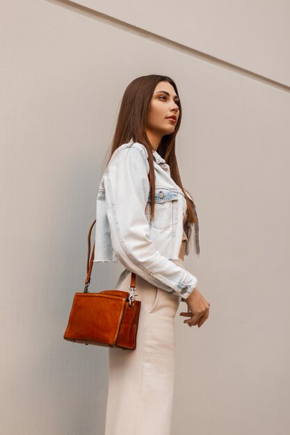 Fashion pretty girl in trendy jeans clothes with bag walks on the street near a modern gray wall