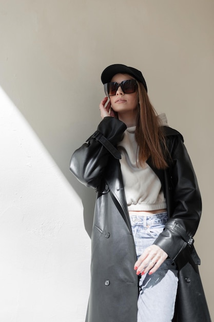 fashion pretty woman hipster with sunglasses and black mock up cap in leather black coat with goodie and jeans stands near a wall on the street