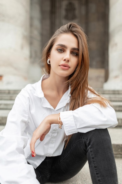 Fashion pretty woman with fresh clean face in casual elegant clothes with white shirt sits near a vintage building in the town