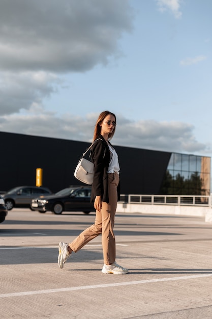 Fashion pretty young woman in vintage casual business clothes with stylish bag in cool sunglasses walks in city parking lot on sunny summer day. Attractive girl in elegant outfit outdoors in city.