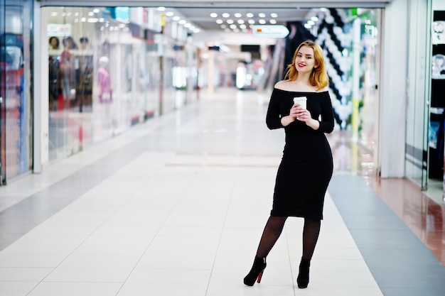 Fashion red haired girl wear on black dress with bright make holding coffee cup at trade shopping center Photo toned style Instagram filters