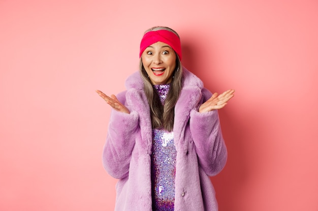 Fashion and shopping concept. Cheerful asian elderly woman in purple stylish coat looking surprised, hear good news and rejoicing, standing over pink background.