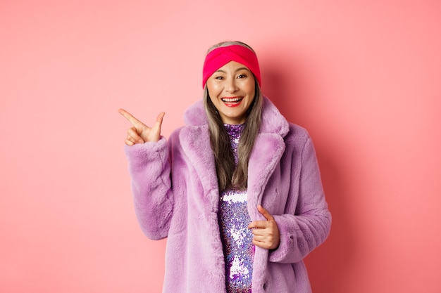 Fashion and shopping concept. Happy asian senior woman checking out trendy promotion, pointing finger left at logo and smiling, wearing winter fake fur coat and headband