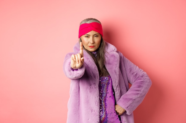 Fashion and shopping concept. Serious asian senior woman showing not so fast gesture, shaking extended finger to stop or warn you, looking determined at camera, wearing stylish purple clothes