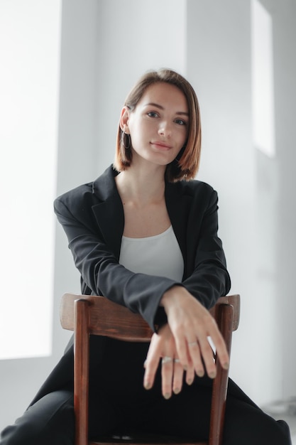 fashion shot of a young stylish woman in a jacket sitting on a chair by the window lifestyle