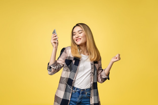 Fashion smiling asian woman listening to music in earphones over yellow background.