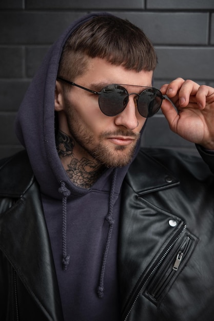 Fashion street male portrait of a handsome business stylish man in fashionable black sunglasses with a hood in a trendy black leather jacket and hoodie on a dark brick background on the street