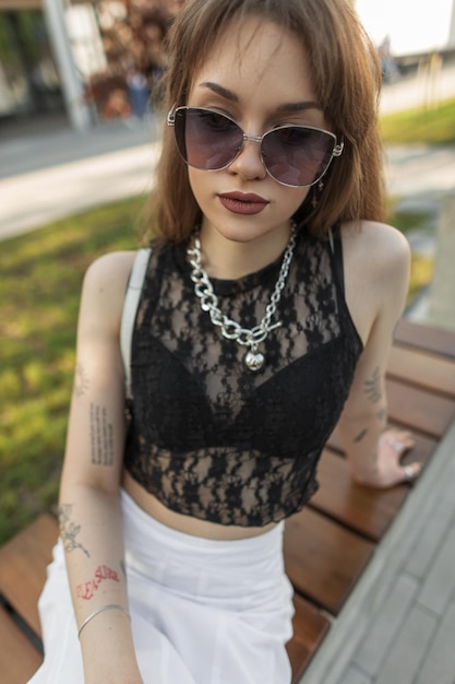 Fashion street sexy woman with vintage sunglasses with jewelry in stylish black top lace and white skirt sits on the bench in the park