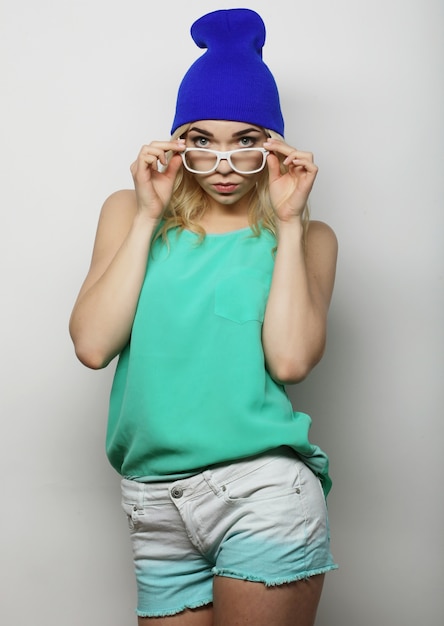 Модный студийный портрет довольно молодой хипстерской блондинки в очках, стильной городской футболке и шляпе, на белом фоне