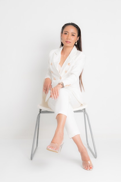 Fashion style catalog clothing for business woman black long hair natural make up wear white suit costume perfect body shape suit sitting on white chair at studio shoot on white background.