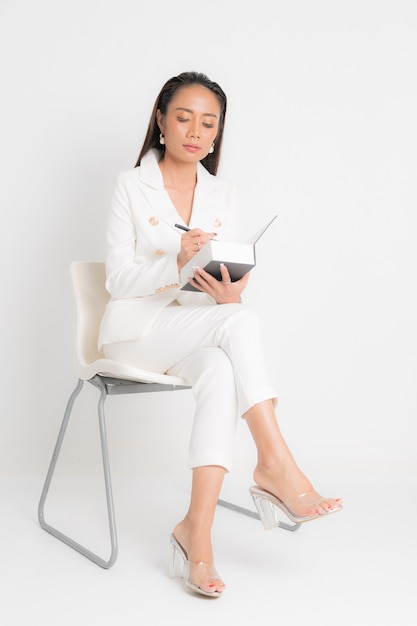 Fashion style catalog clothing for business woman black long hair natural make up wear white suit sitting on white chair, holding book and pens at studio shoot on white background.