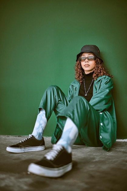 Photo fashion style and clothes with a model black woman sitting on the floor against a green background portrait glasses and fashionable with a young female posing to promote trendy or edgy clothing